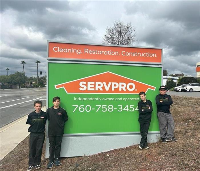 Four young team members stand in front of a large SERVPRO sign, wearing black SERVPRO uniforms and smiling confidently.